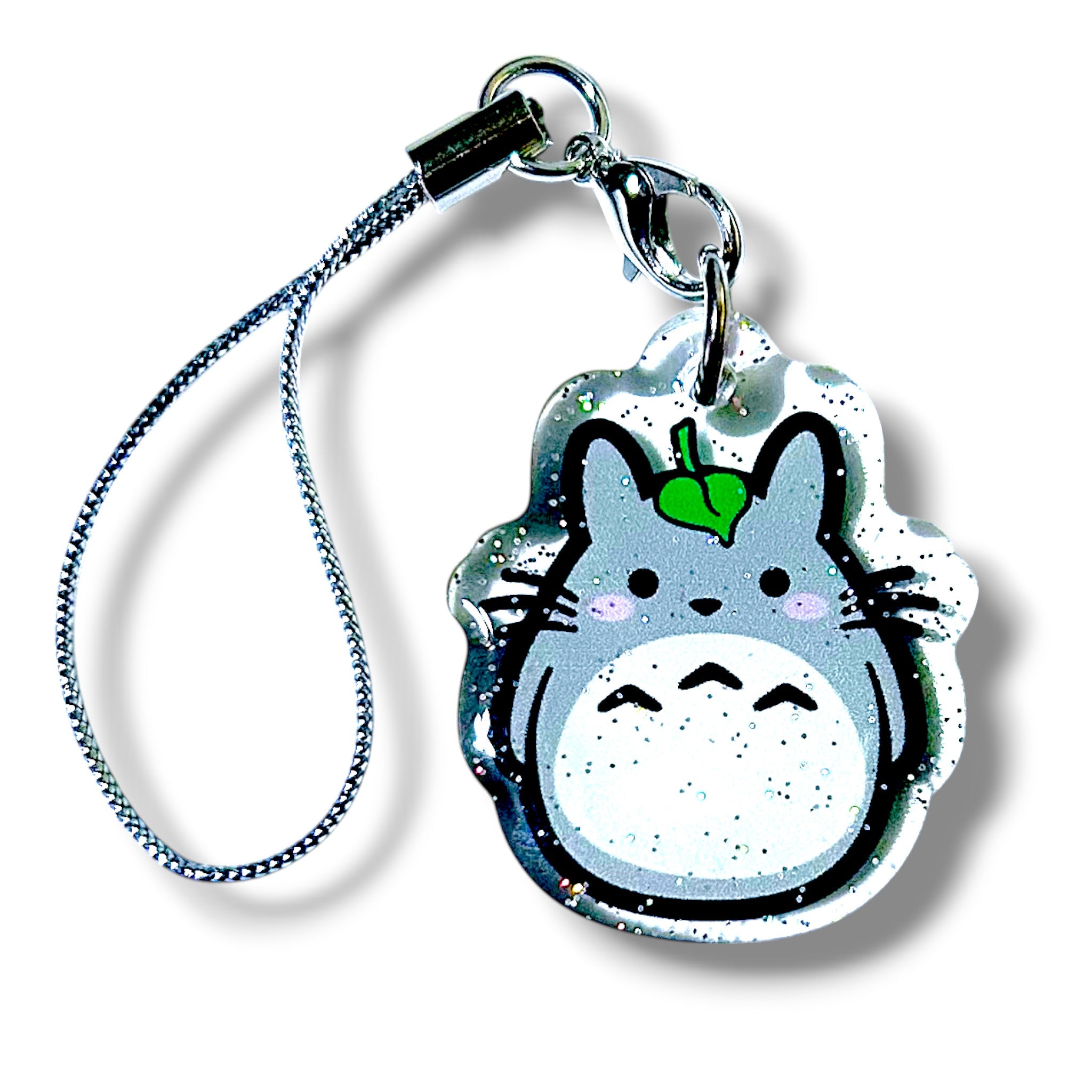 Glossy acrylic keychain charm featuring a round forest creature with gray fur, a white belly marked by three black ovals, and a tan face. The character has small ears, wide eyes, and a tiny green leaf resting on its head. Embedded glitter adds sparkle throughout the charm. It is attached to a silver strap with a metal clasp and ring. The design blends whimsical forest aesthetics with a magical, anime-inspired style, making it a charming collectible accessory.