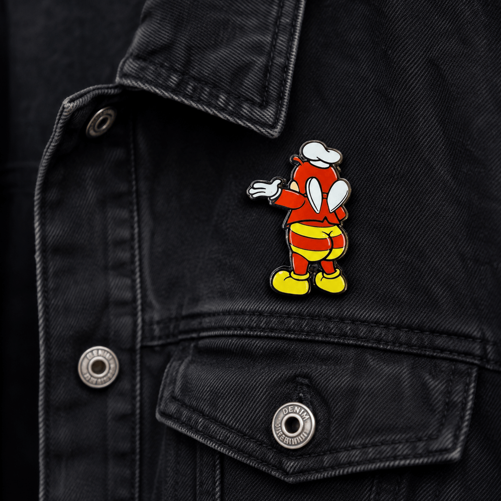 Jolli-Butt enamel pin attached to a black denim jacket, featuring a red character in a white chef’s hat, striped shorts, and yellow boots.
