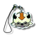 Cartoon-style keychain charm featuring a stylized fluffy creature with a white face, closed eyes, a protruding tongue, and a bold orange arrow on its forehead. The character has a rounded body and a soft, relaxed expression. It is attached to a black strap with a silver clasp. The design blends playful, air-themed aesthetics with cozy, animal-like features, making it a whimsical and collectible accessory.