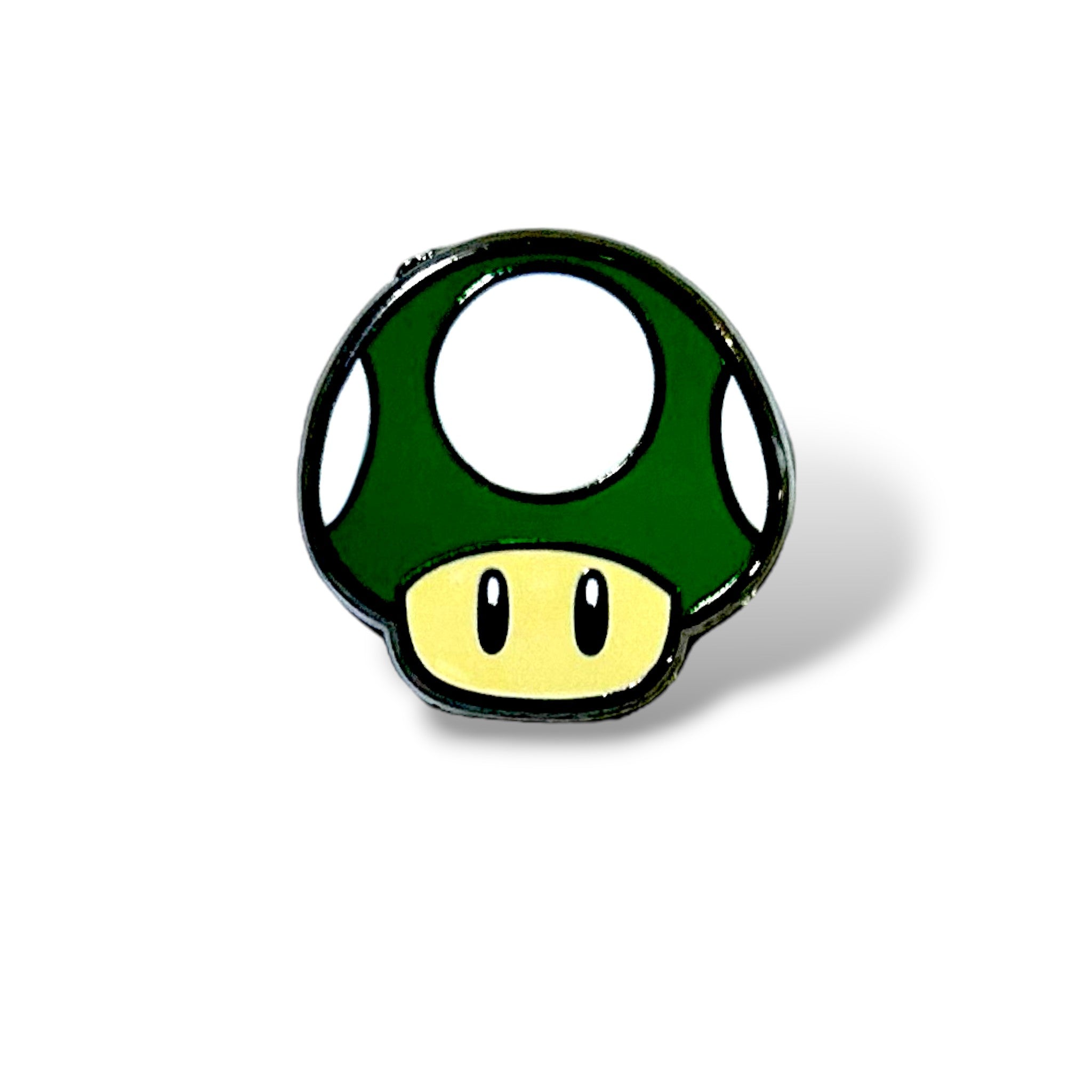 Enamel pin featuring a green mushroom with a beige stem, white spots on the cap, and two black oval eyes; designed in a cartoon style inspired by classic gaming power-ups, with a glossy finish and bold outline.