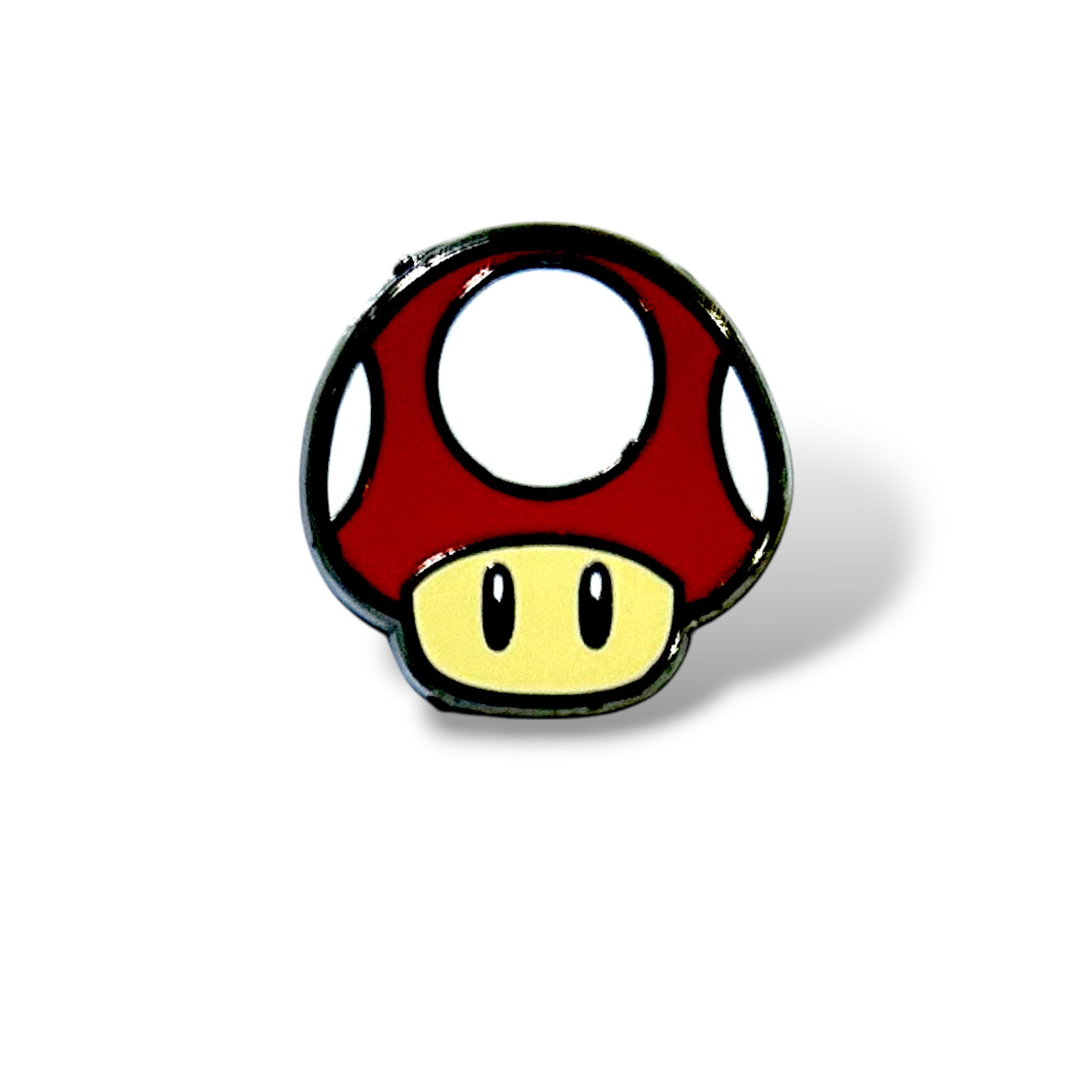 Enamel pin shaped like a red mushroom with a beige stem, white spots on the cap, and two black oval eyes; designed in a cartoon style inspired by classic gaming power-ups, with a glossy finish and bold outline.
