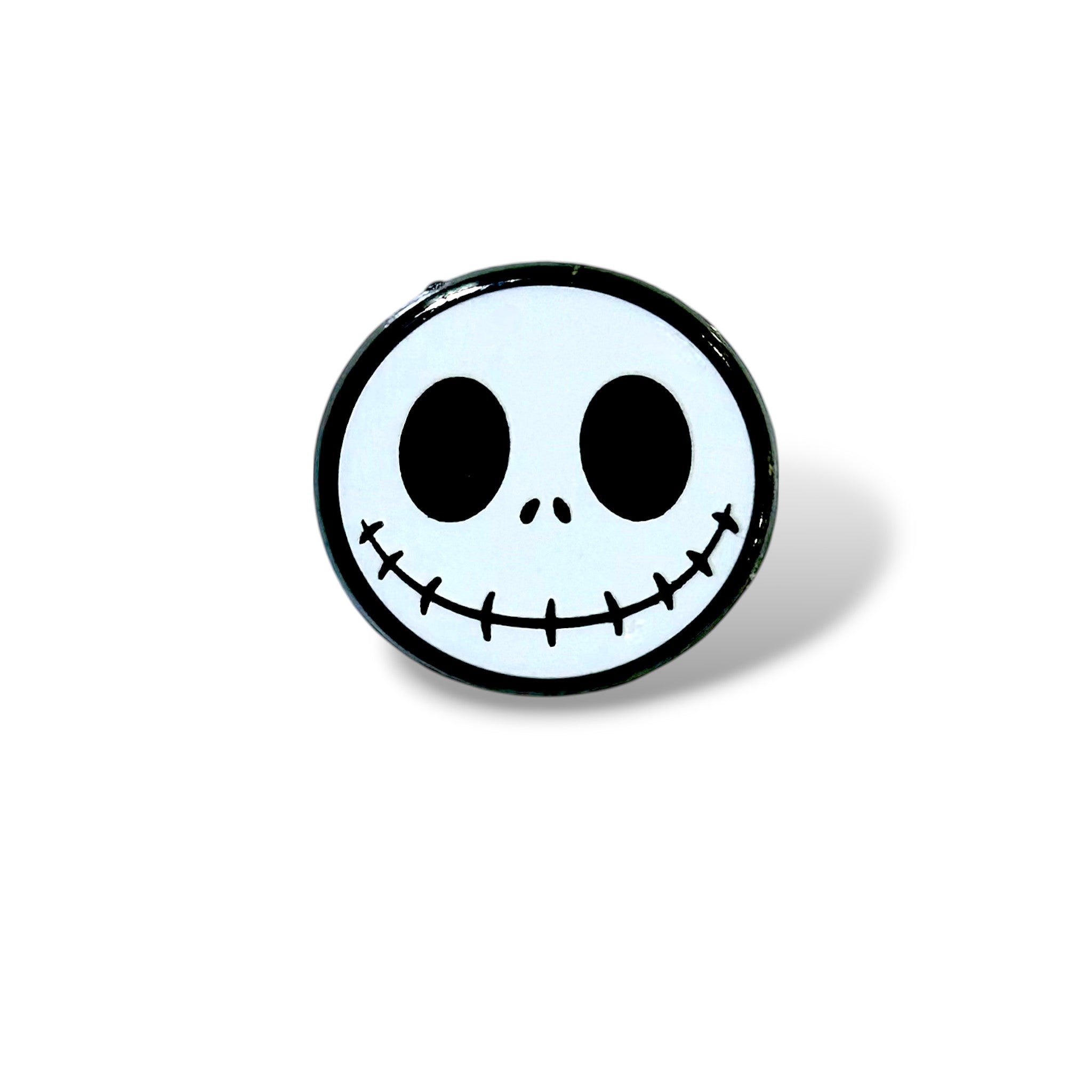 Enamel pin featuring a stylized white skull face with large black oval eyes, two small nostrils, and a wide stitched smile; inspired by a spooky cartoon character and designed in a clean, minimalist Halloween style with bold black outlines.